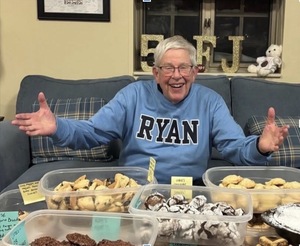 Fr. Joe Carey, known as FJ. A smiling older man in a light blue "RYAN" shirt proudly displays clear containers of various homemade cookies.