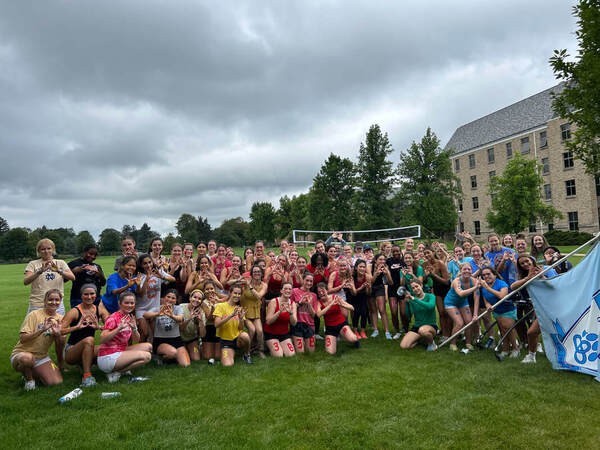 Image of Ryan Girls after Section Olympics in their respective section colors. Ryan flag is also featured. Girls are posing outside Ryan Hall on McGlinn Fields.